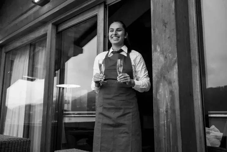 Smiling chalet host in apron holding two champagne flutes on wooden terrace, offering personalised service