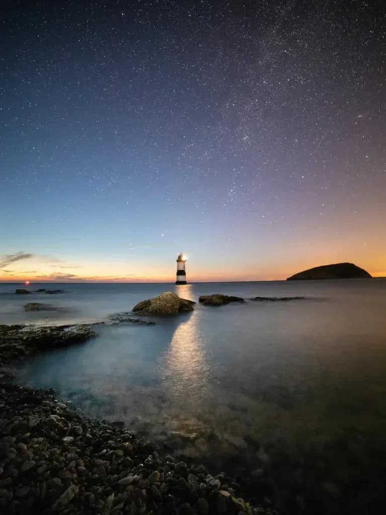 Trwyn Du lighthouse under starry night sky with Milky Way visible, reflecting on calm Welsh waters at twilight