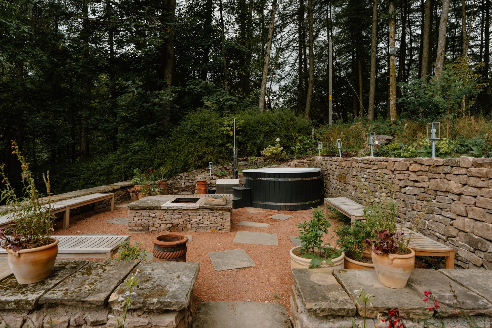 Secluded woodland terrace with a wood-fired hot tub, stone fire pit, and rustic dry-stone walls at a luxury Peak District ho…