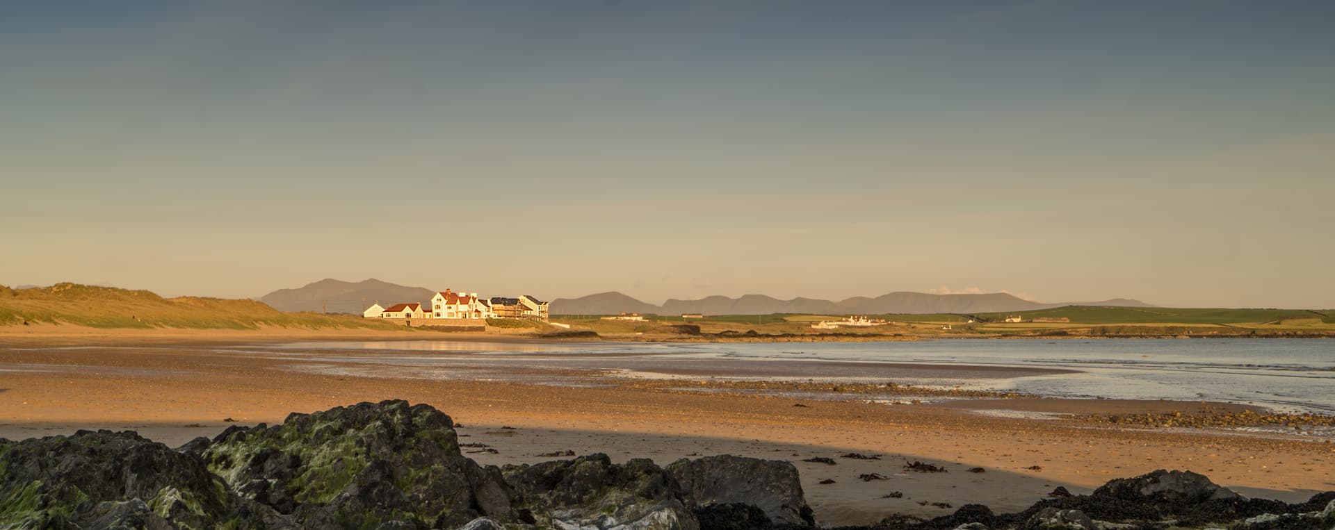 Golden hour over a wide sandy beach on Anglesey with coastal cottages and Snowdonia mountains in the distance