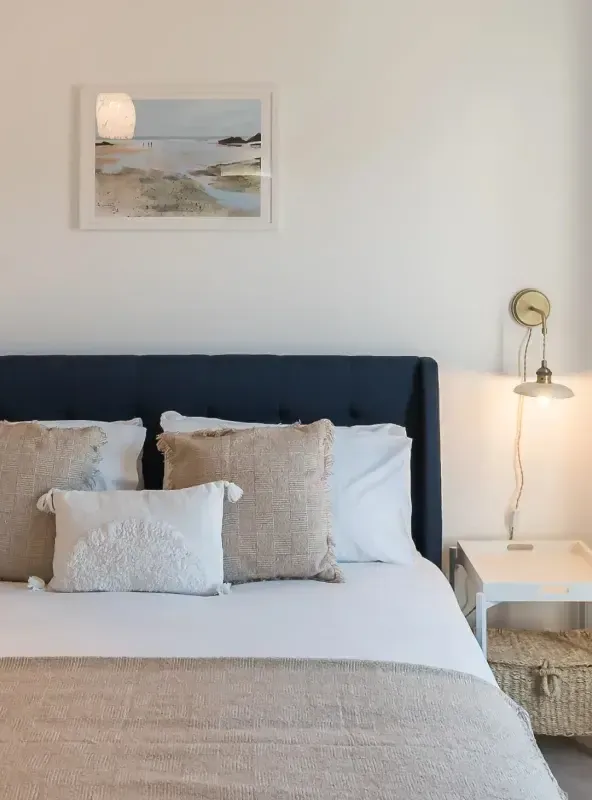 Coastal holiday let bedroom with navy buttoned headboard, layered neutral cushions, brass wall lamp, and framed beach artwor…