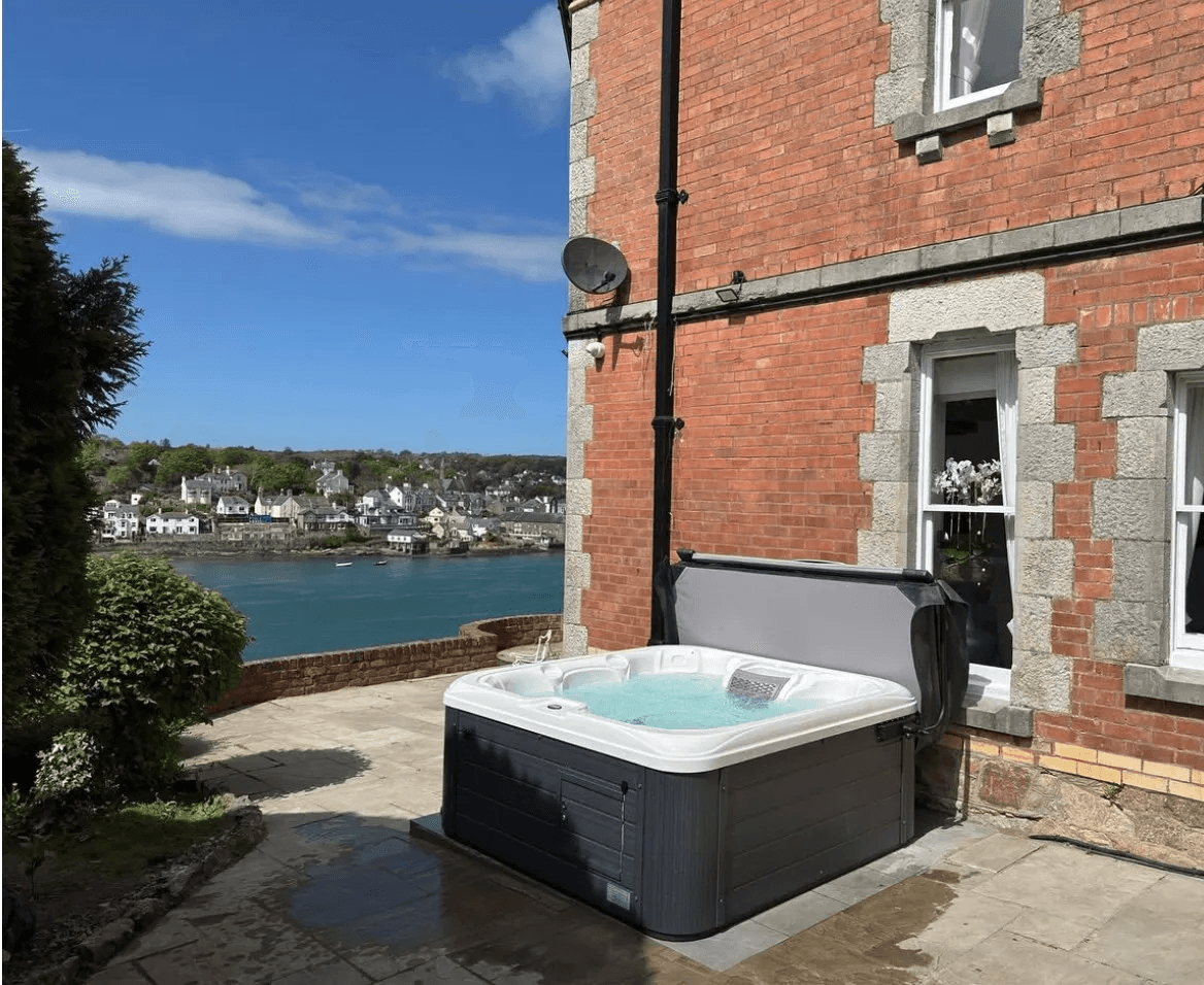 Outdoor hot tub on a stone terrace beside a red brick holiday cottage, overlooking a turquoise estuary and coastal village