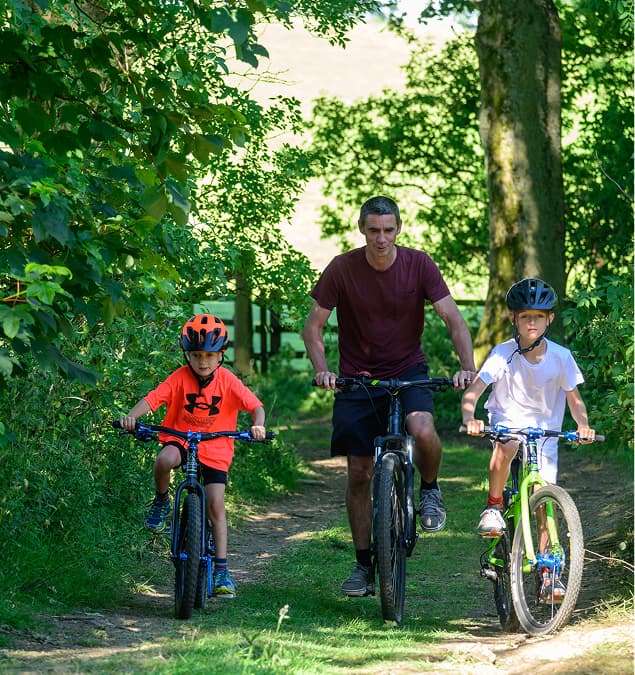 Father and two children cycling together on woodland path surrounded by lush green trees on sunny day