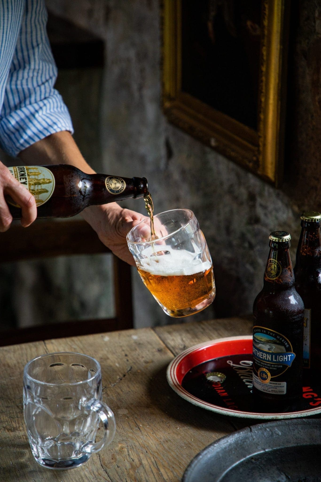 Orkney ale being poured into a glass tankard at a rustic pub table, with Northern Light bottles on a vintage serving tray ne…