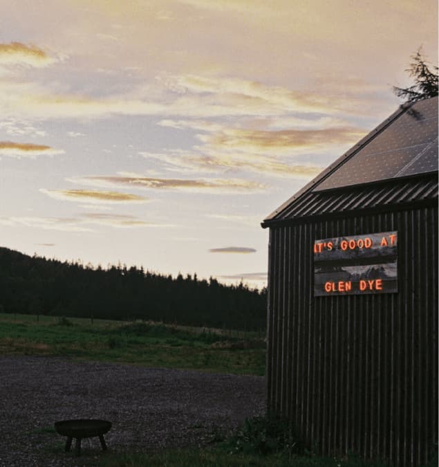 Glowing neon sign reading It's Good at Glen Dye on a dark timber clad barn at dusk, with a fire pit and Scottish woodland be…