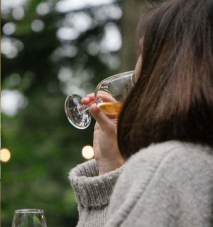 Guest in a cosy knit jumper sipping white wine outdoors, surrounded by soft bokeh lights and lush greenery