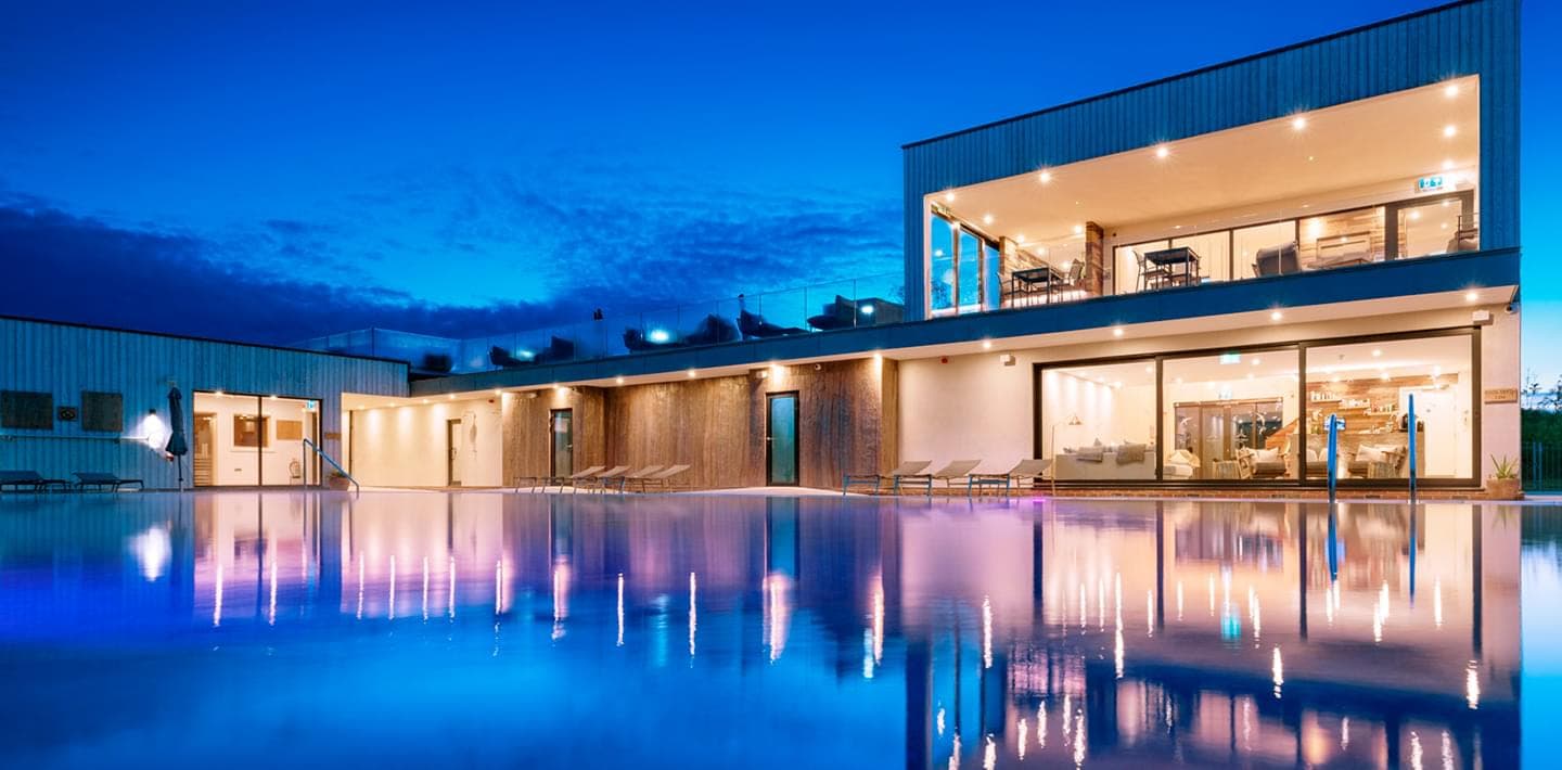 Floodlit outdoor pool at dusk beside a modern two-storey holiday let with floor-to-ceiling glazing and sun loungers