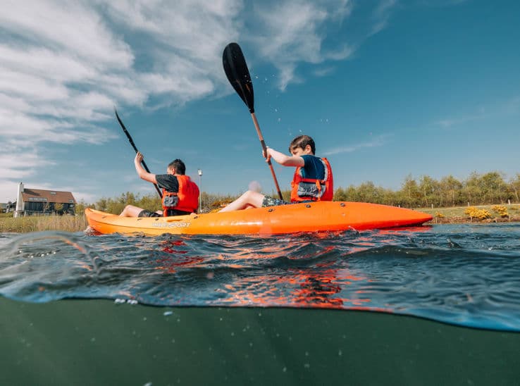 Two guests paddling an orange tandem kayak on the calm lake at Silverlake holiday park, shot from water level on a sunny day