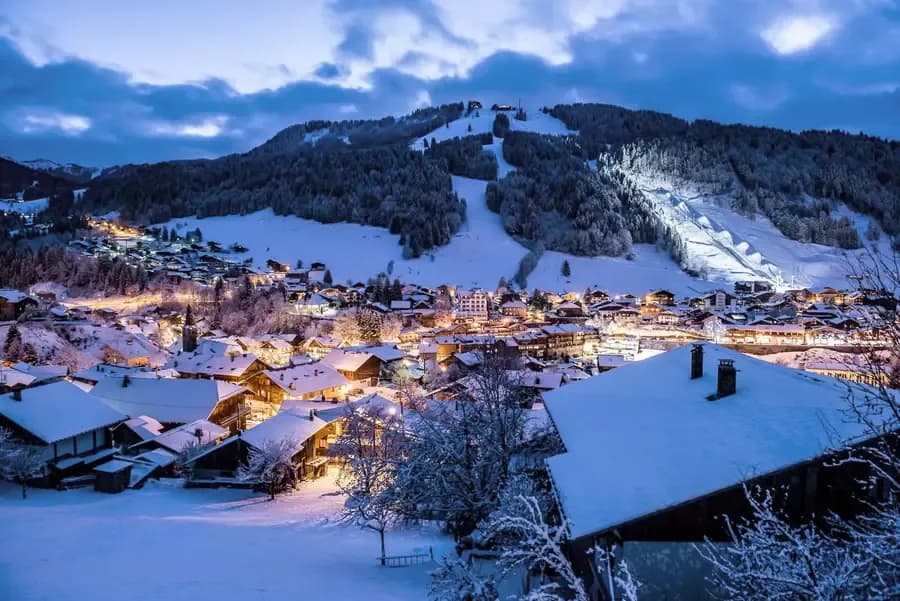 Snow-covered Alpine village at dusk with illuminated chalets and ski slopes, perfect for luxury winter holiday lets