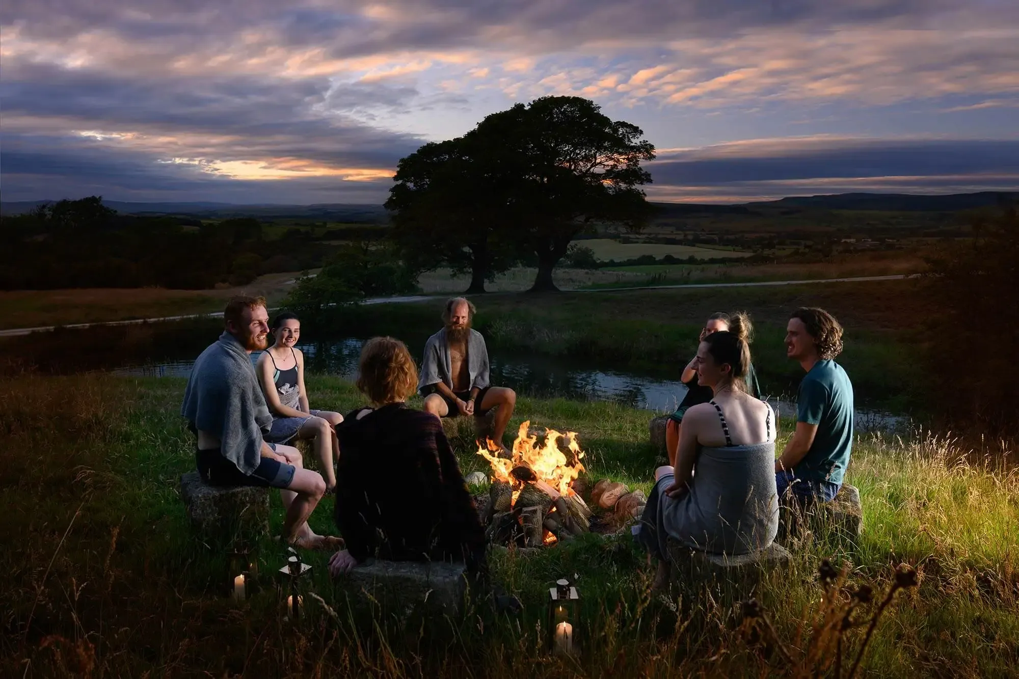 Group gathered around crackling fire pit at dusk with rolling countryside views and ancient oak tree