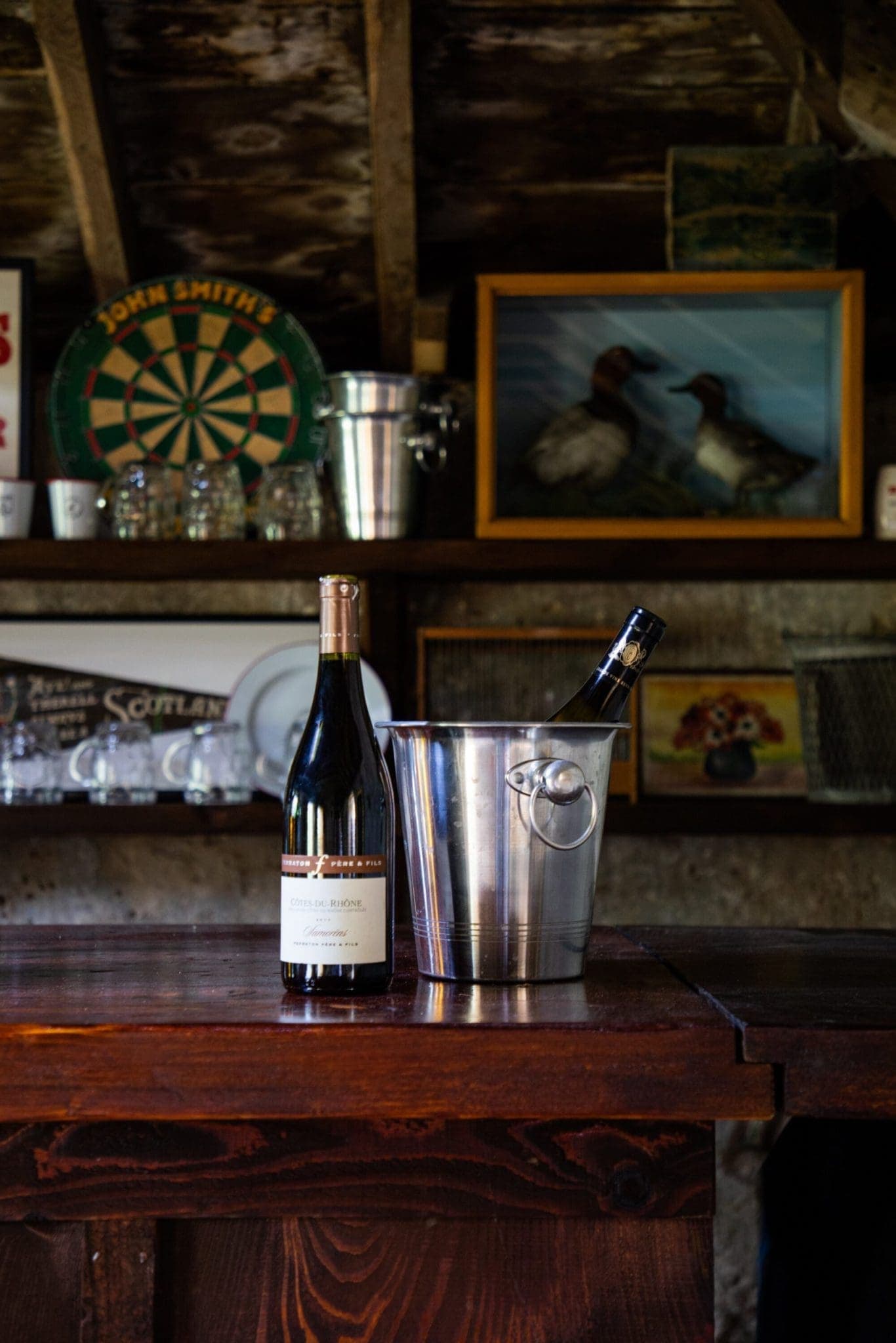 Rustic games room bar with a Côtes du Rhône bottle and champagne ice bucket on a dark wood counter, dartboard and vintage Sc…