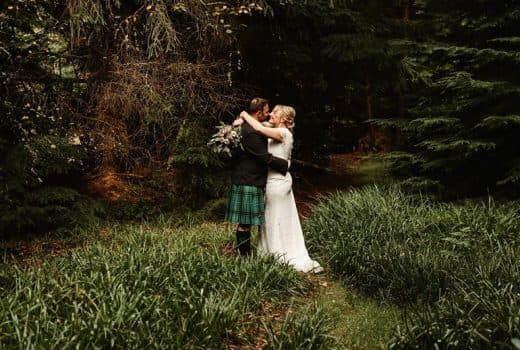 Bride and groom embracing in a lush woodland clearing, groom wearing a traditional Scottish tartan kilt