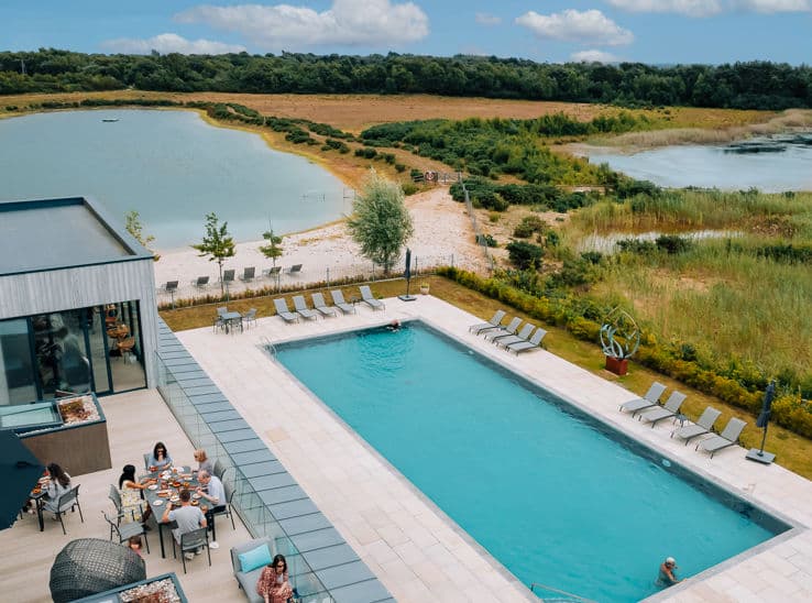 Aerial view of a large outdoor pool with sun loungers beside a private lake and sandy beach at a luxury holiday let in the C…