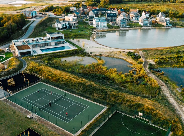 Aerial view of a luxury holiday estate with a full-size tennis court, outdoor pool, and lakeside properties set among open c…