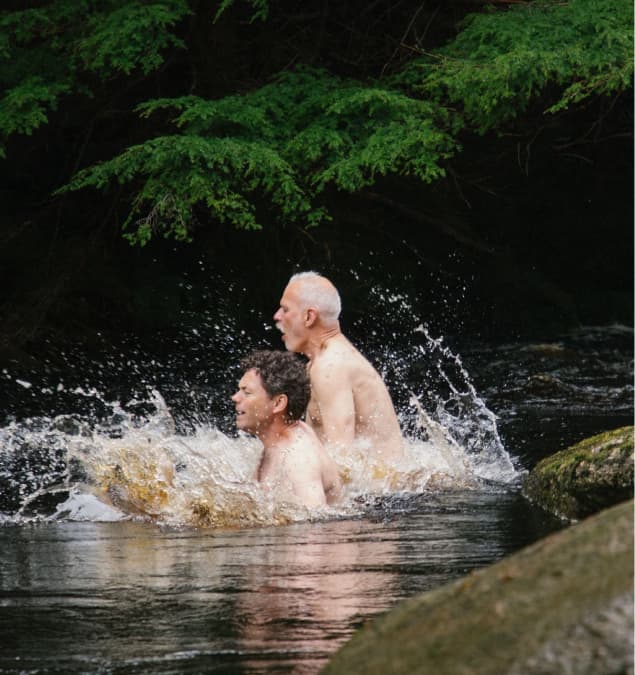 Two guests wild swimming in a rocky woodland river, splashing in clear water beneath lush green trees