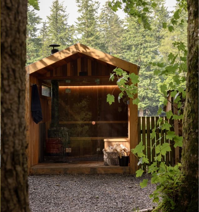 Timber outdoor sauna cabin with glass front, wood store, and flue pipe nestled among tall woodland trees