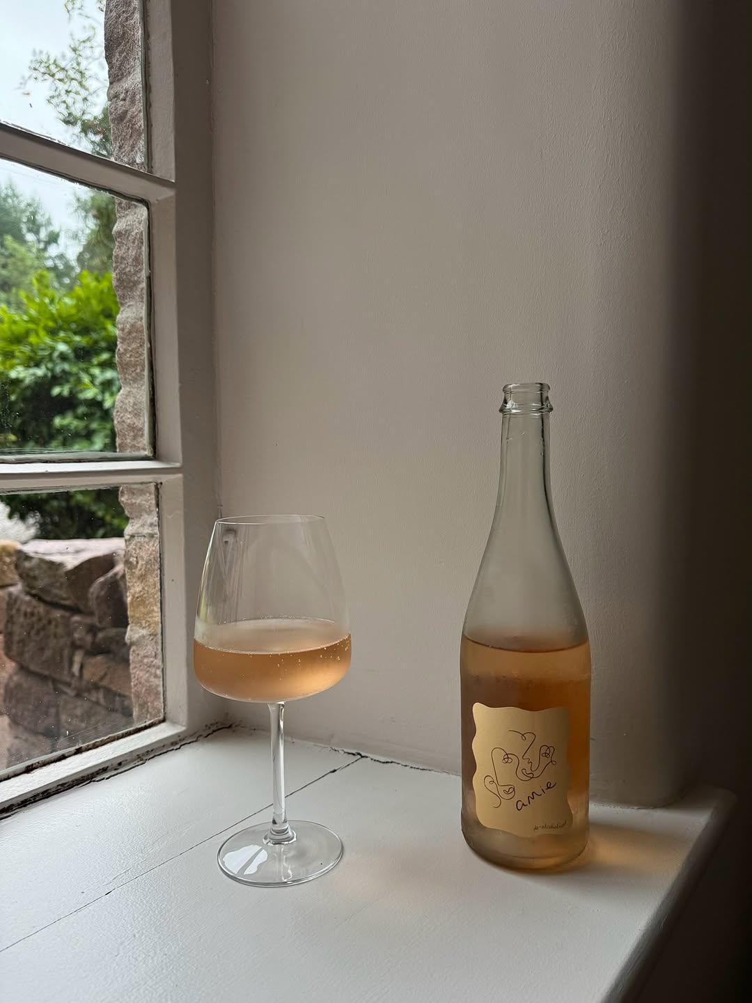Glass of sparkling rosé wine beside an Amie natural wine bottle on a stone cottage windowsill, with lush garden greenery bey…