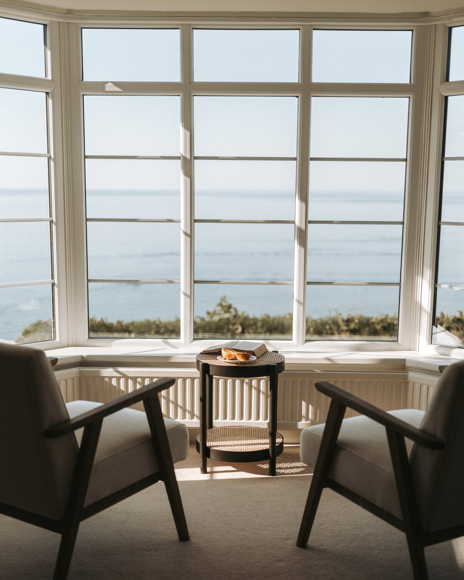 Sunlit bay window sitting room with two armchairs and a cane side table overlooking a calm coastal sea view