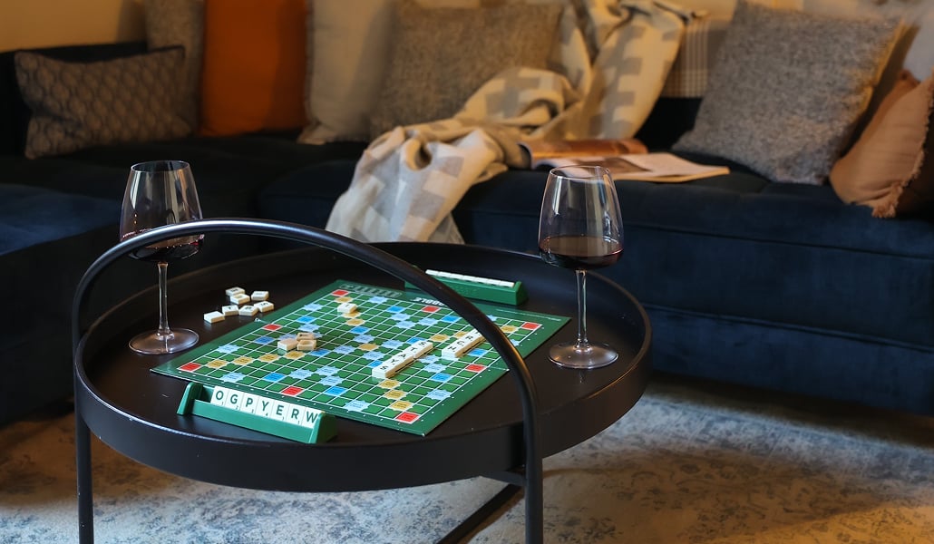 Scrabble board and two glasses of red wine on a round coffee table in a cosy holiday let snug with a navy velvet sofa and so…