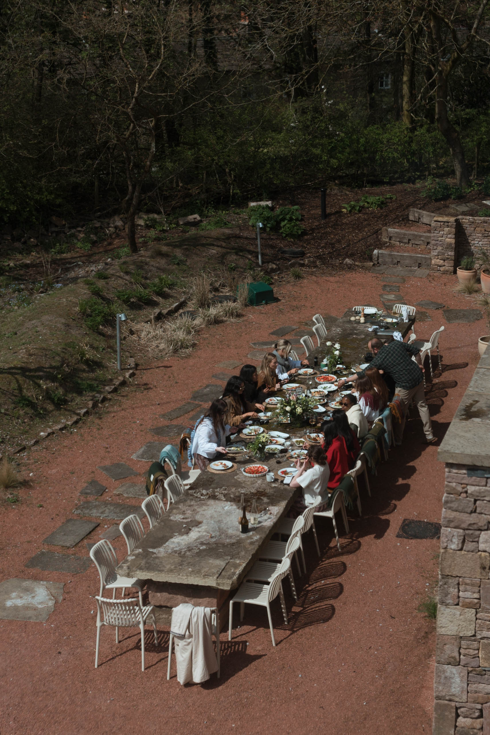 Large group of guests dining al fresco at a long stone table on a sunlit terrace surrounded by woodland, sharing a feast dur…