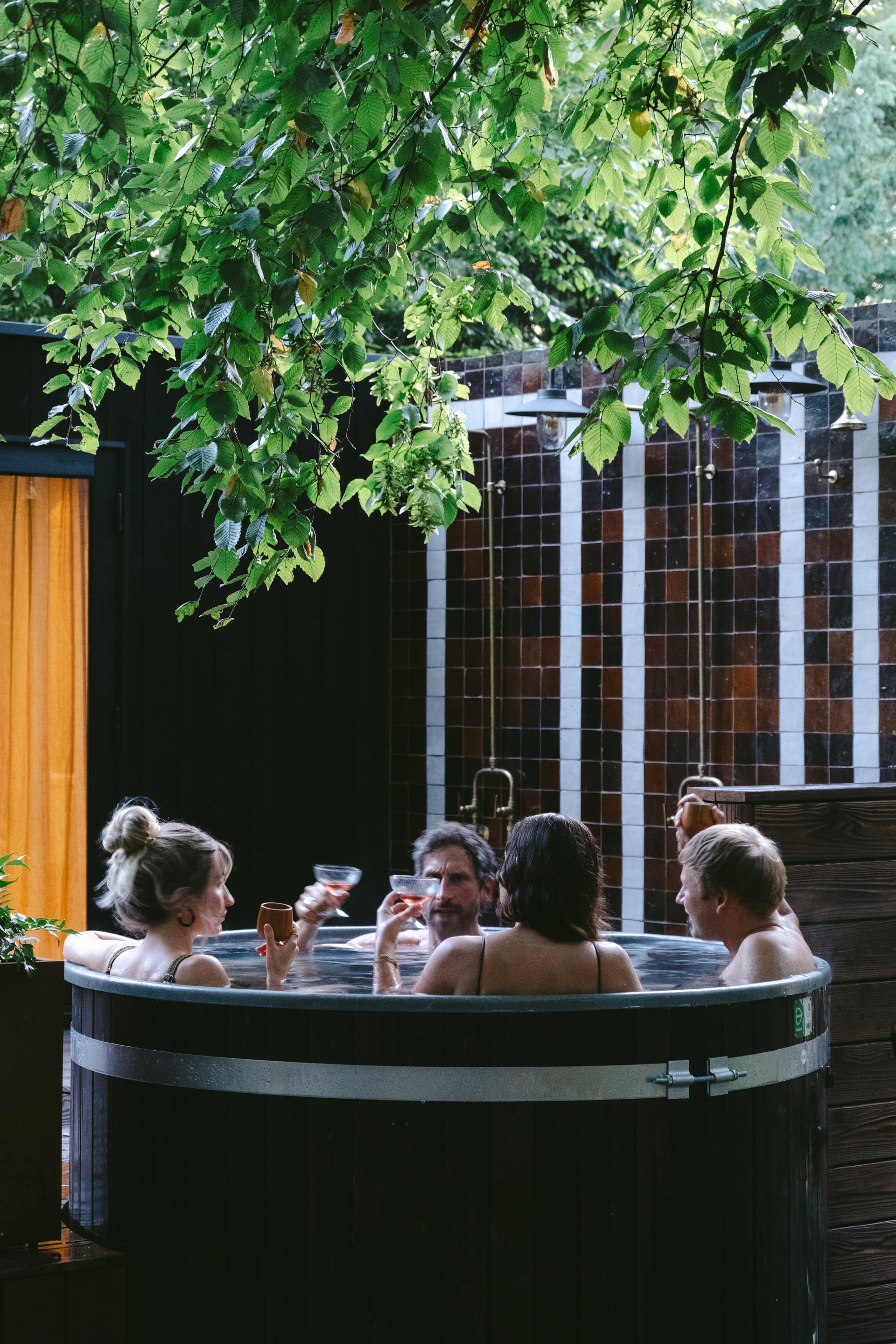 Four guests relaxing in an outdoor wooden hot tub with drinks, surrounded by lush green tree canopy and dark tiled shower wa…