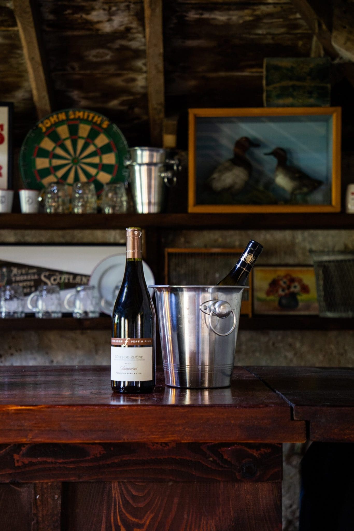 Rustic games room bar with a Côtes du Rhône bottle and champagne ice bucket on a dark wood counter, dartboard and vintage Sc…
