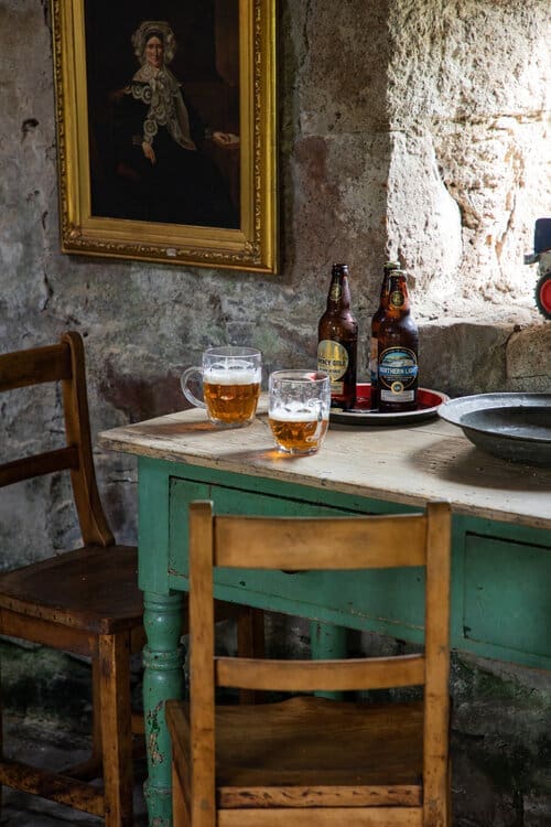 Rustic pub corner with two pints of ale, bottled beers, and a vintage oil portrait on a weathered stone wall
