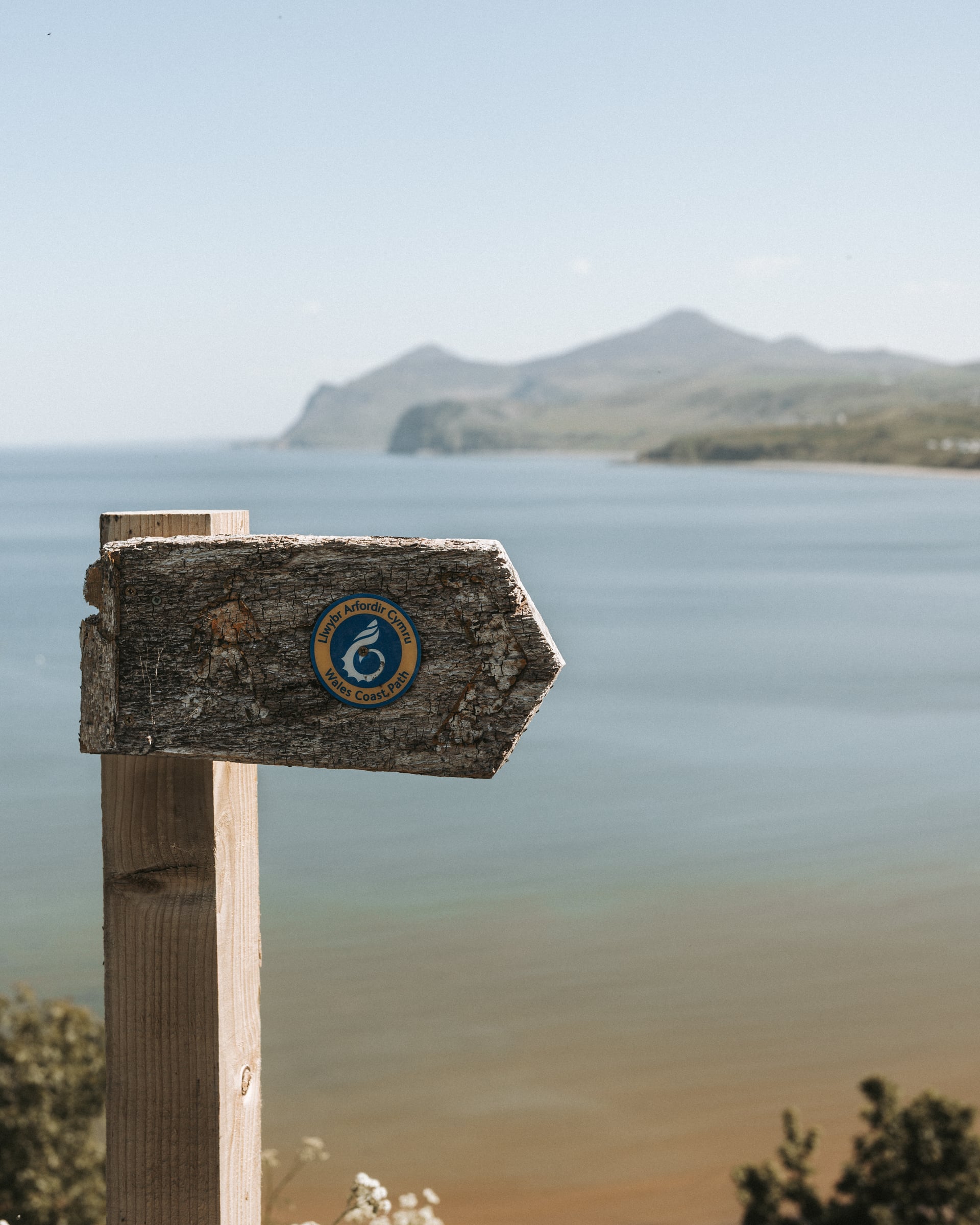 Wales Coast Path wooden waymarker sign with calm bay and headland stretching into the distance on a clear day