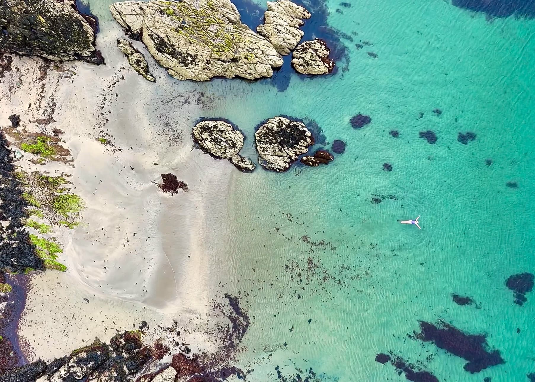 Aerial view of a lone swimmer floating in crystal-clear turquoise water beside a secluded white sand beach with rocky outcro…