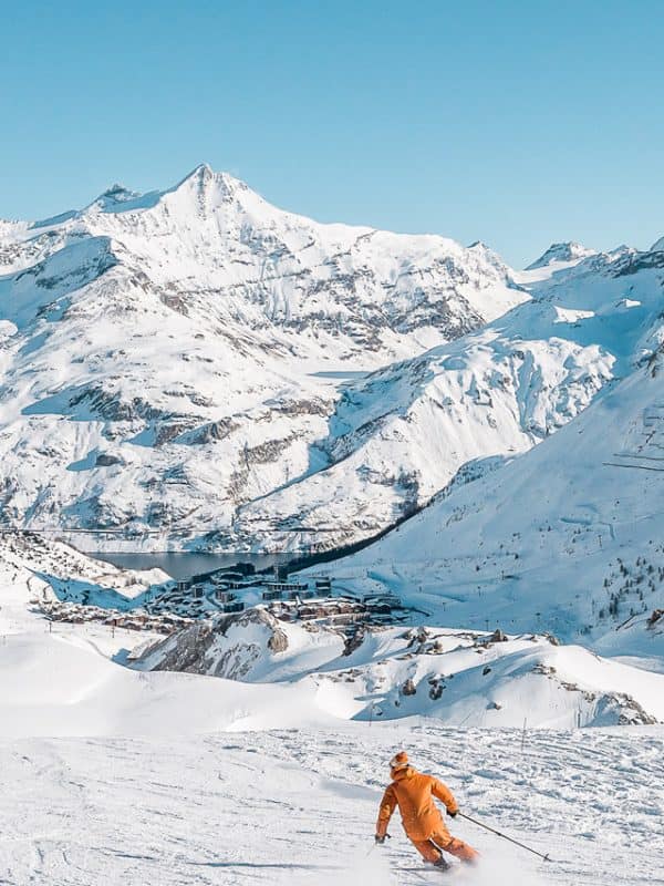 Skier in orange jacket carving down pristine slopes with dramatic Alpine peaks and luxury ski resort village below