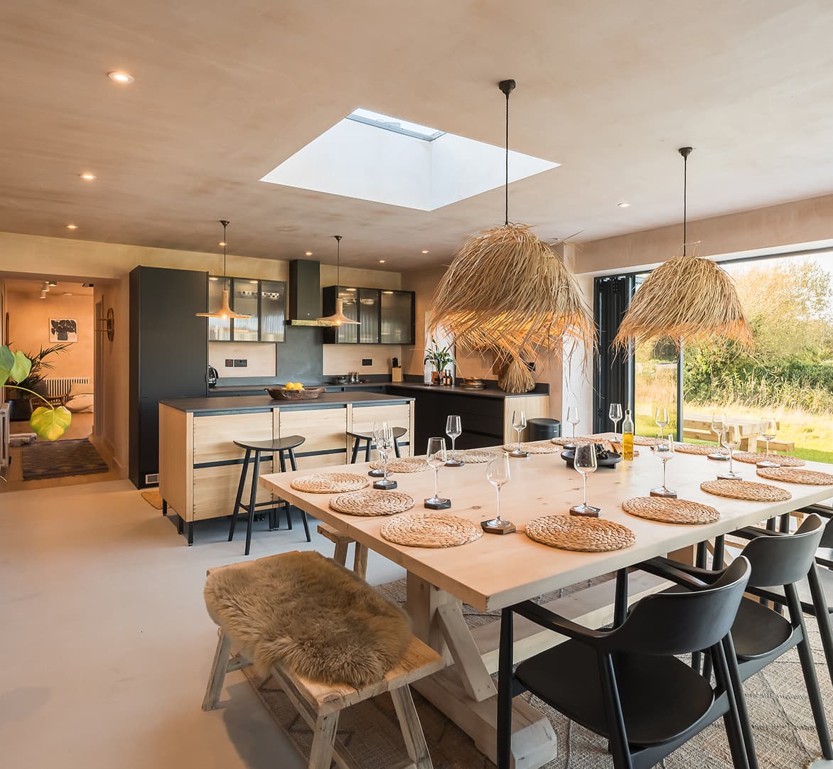 Open-plan kitchen and dining area with large wooden table set for ten, rattan pendant lights, skylight, and bi-fold doors op…