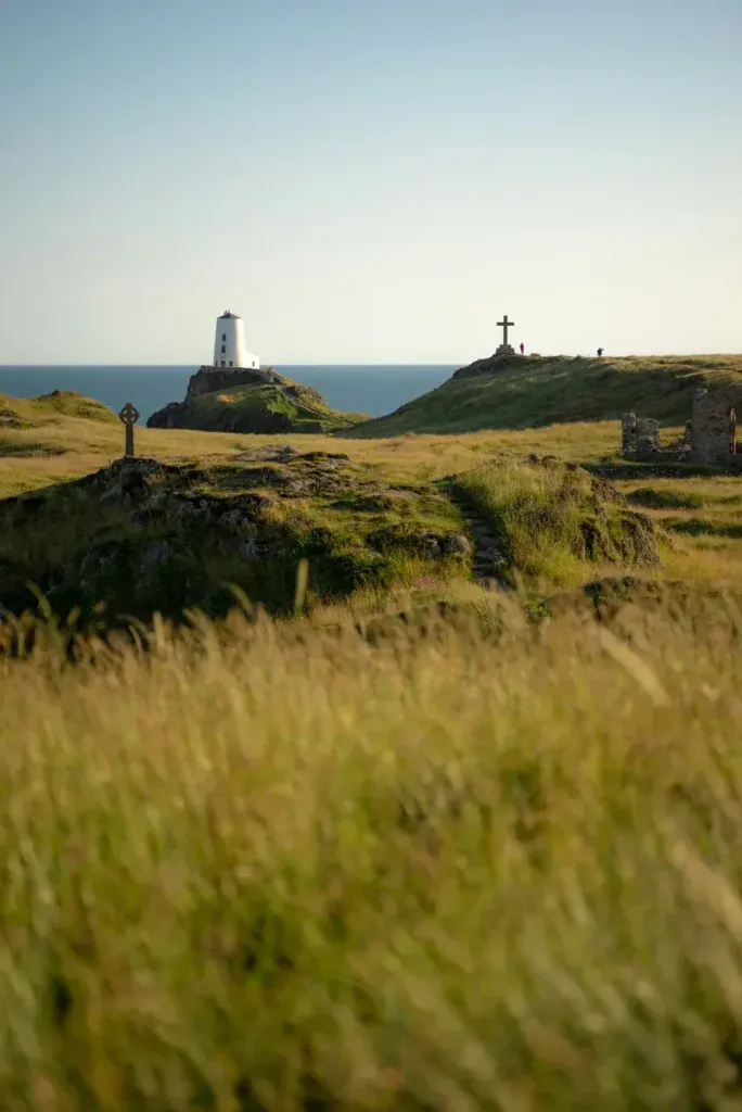 White lighthouse and memorial cross on grassy clifftops at Llanddwyn Island, Anglesey, with golden meadows and sea views