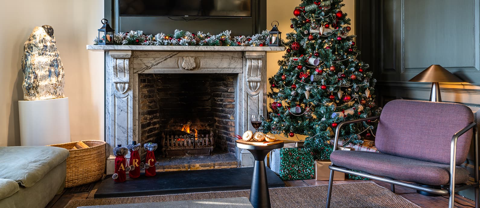 Elegant sitting room dressed for Christmas with a lit marble fireplace, decorated tree, mince pies and red wine beside wrapp…