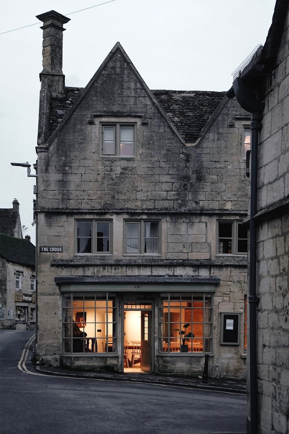 Honey-stone Cotswold building at dusk with warm light glowing from the ARC shopfront at The Cross in Painswick