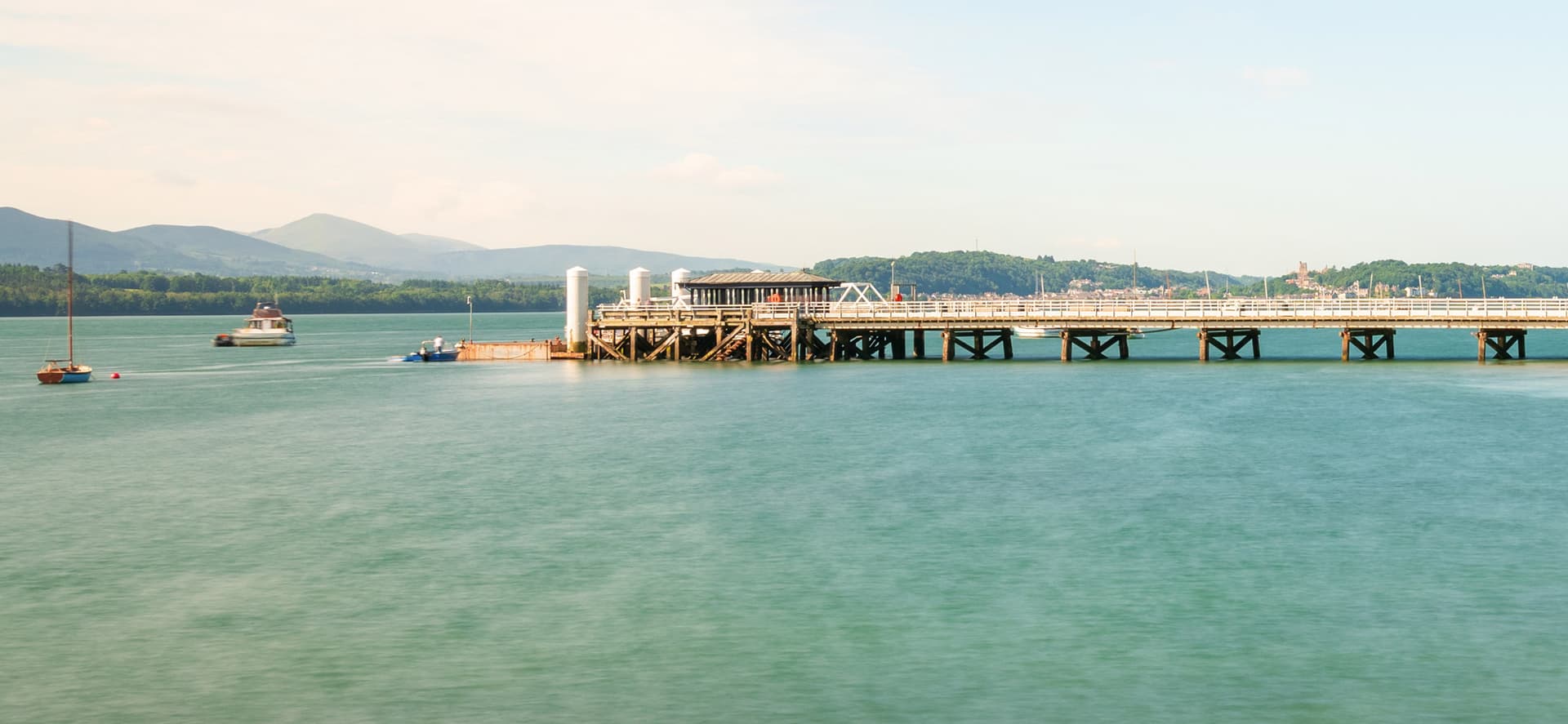 Victorian pier stretching over calm turquoise waters with wooded hills and mountains beyond, on the North Wales coastline