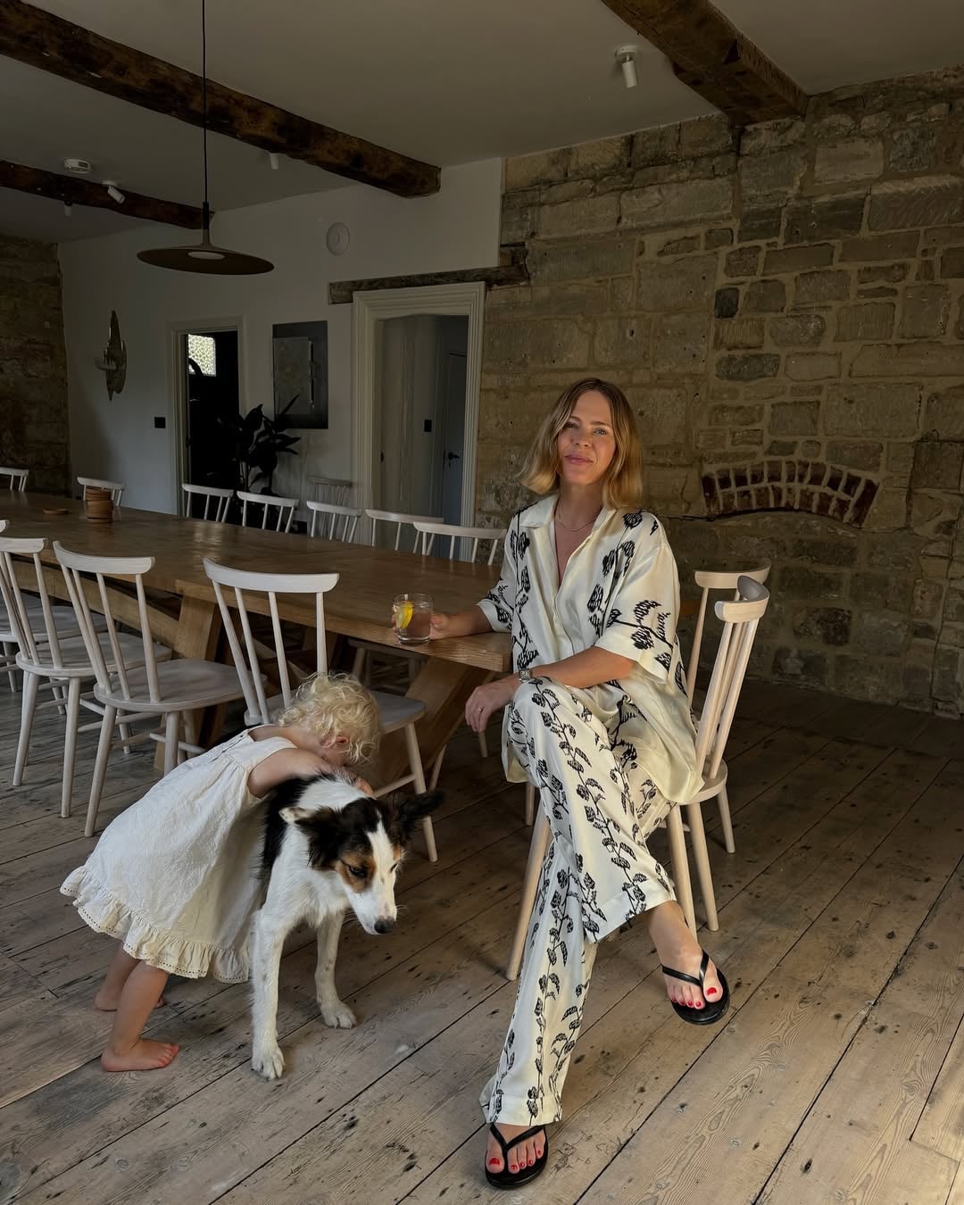 Woman relaxing at a long oak dining table in a stone-walled barn conversion, with a young child and border collie dog on rus…