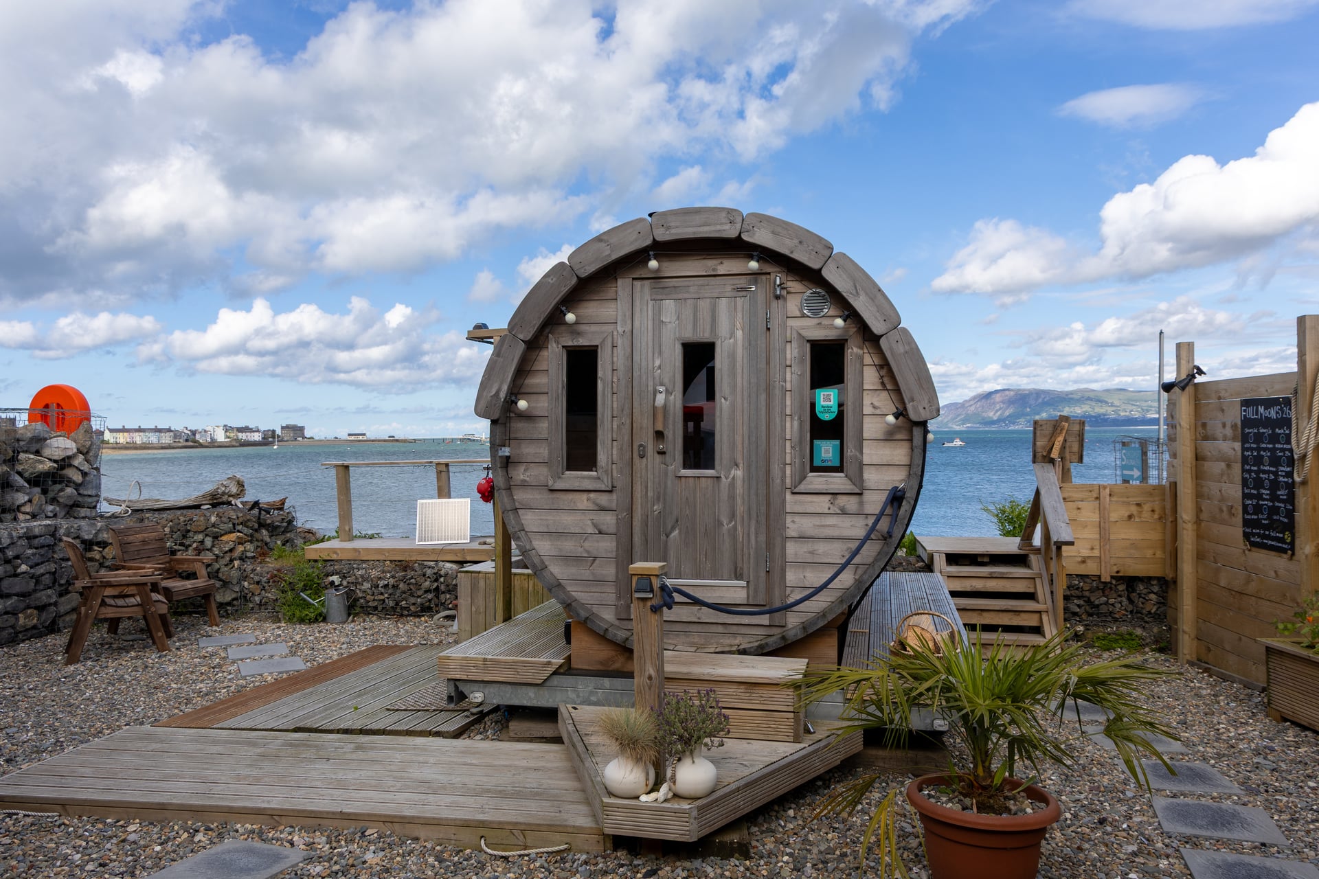 Barrel sauna on a timber deck beside the Welsh coastline, with mountain and bay views stretching behind it