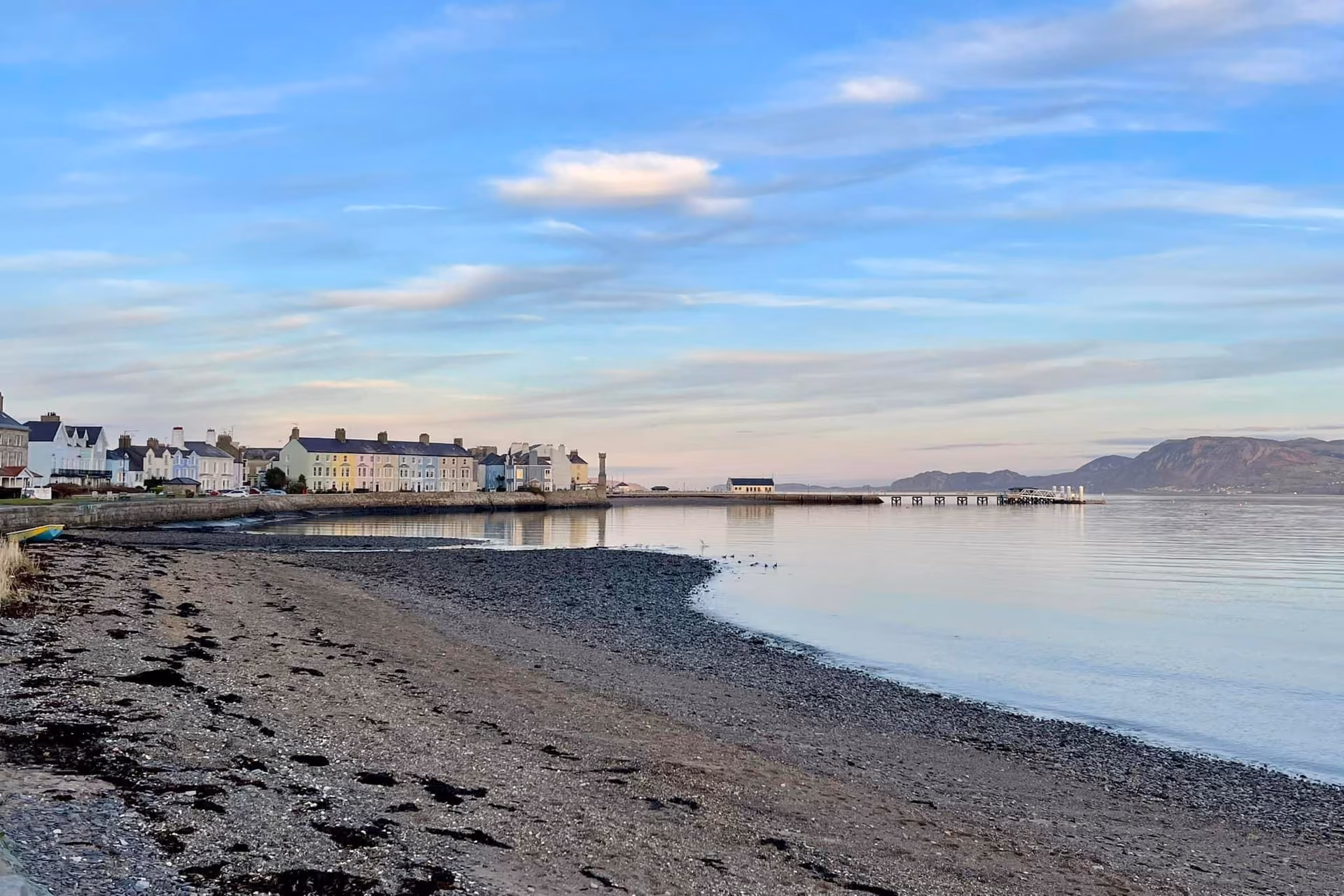 Calm shingle shoreline at Beaumaris on Anglesey, with pastel-fronted Georgian townhouses lining the seafront and mountains b…