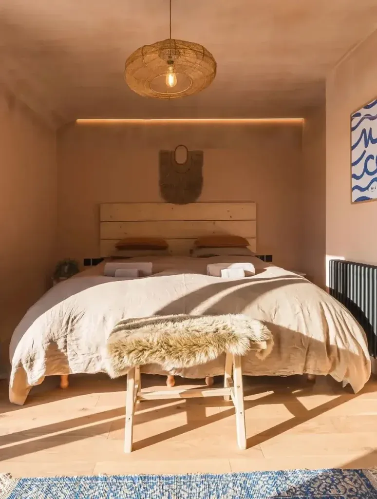 Sun-drenched double bedroom with linen bedding, sheepskin throw, pine headboard, and woven rattan pendant light in a warm-to…