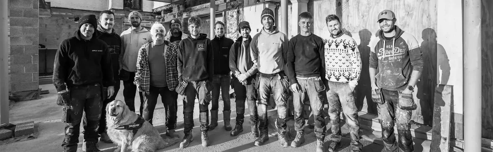 Birch Stays construction team posing together inside a property mid-renovation, with a golden retriever, captured in black a…