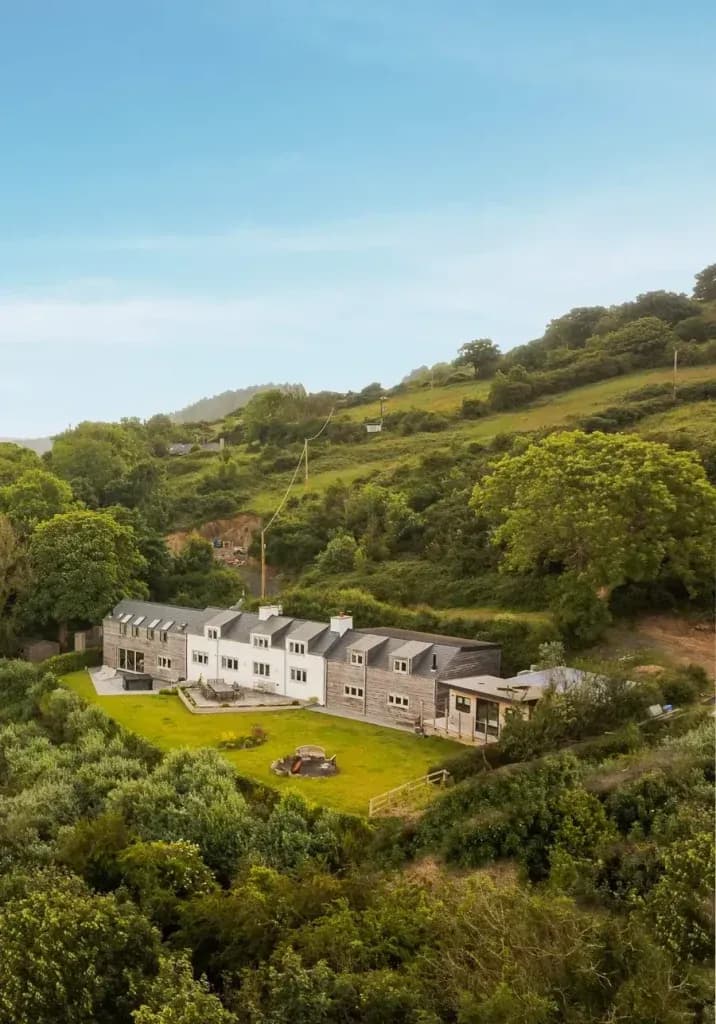 Aerial view of a large rural holiday cottage nestled in a lush green valley, with a lawned garden and fire pit seating area