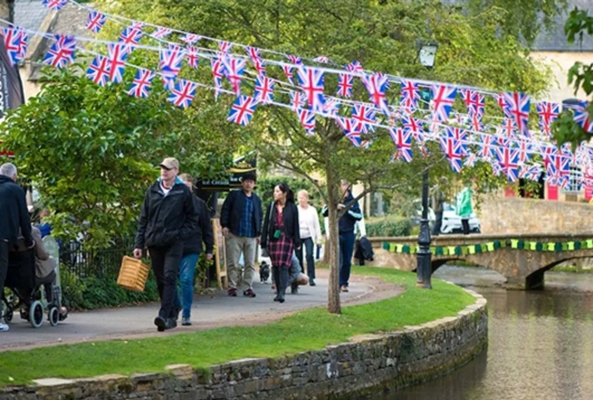 Visitors strolling along the river in Bourton-on-the-Water, Cotswolds, with Union Jack bunting strung between trees during a…