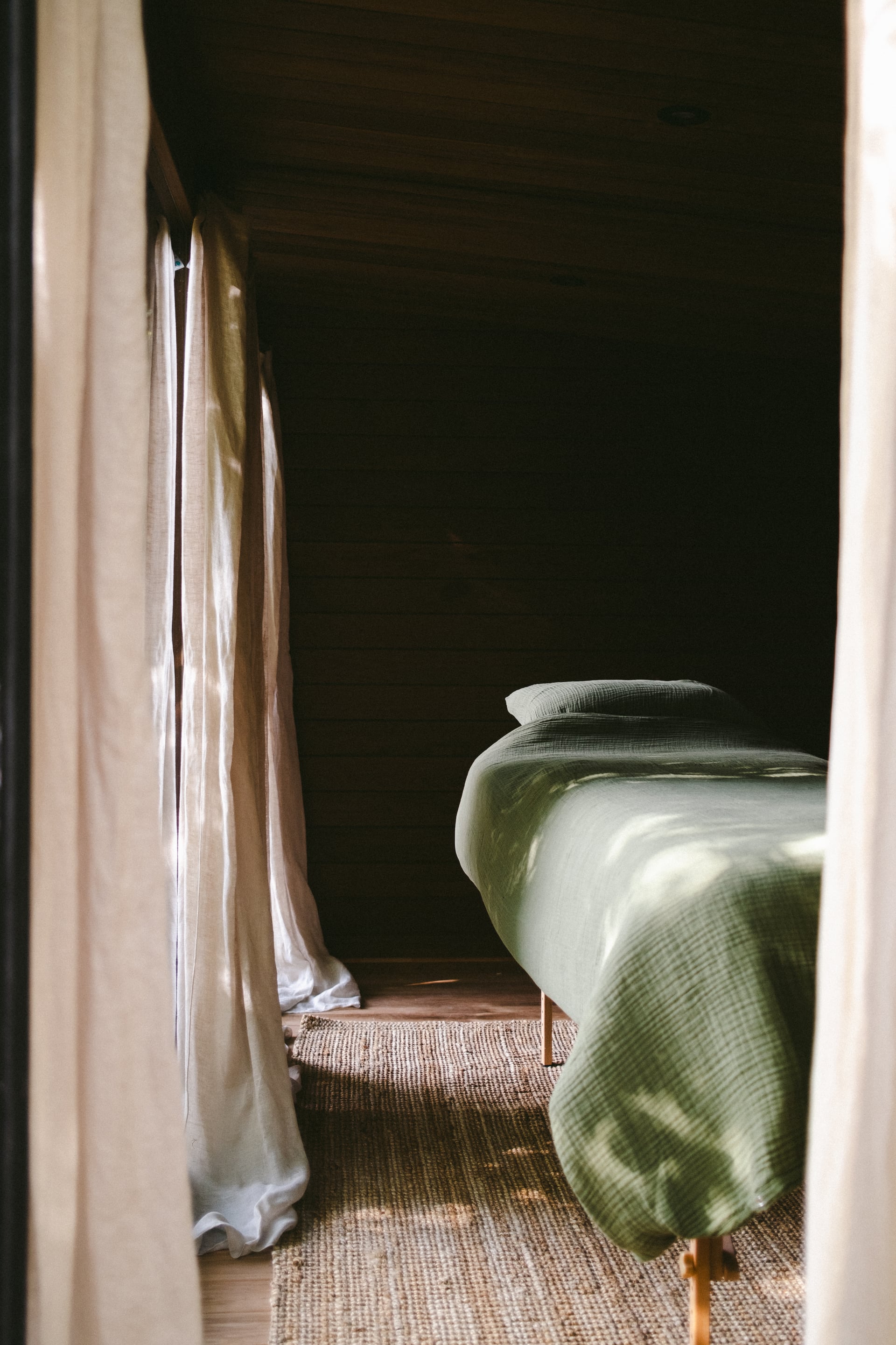Cosy cabin bedroom with sage green linen bedding, dark timber walls, and soft natural light filtering through linen curtains