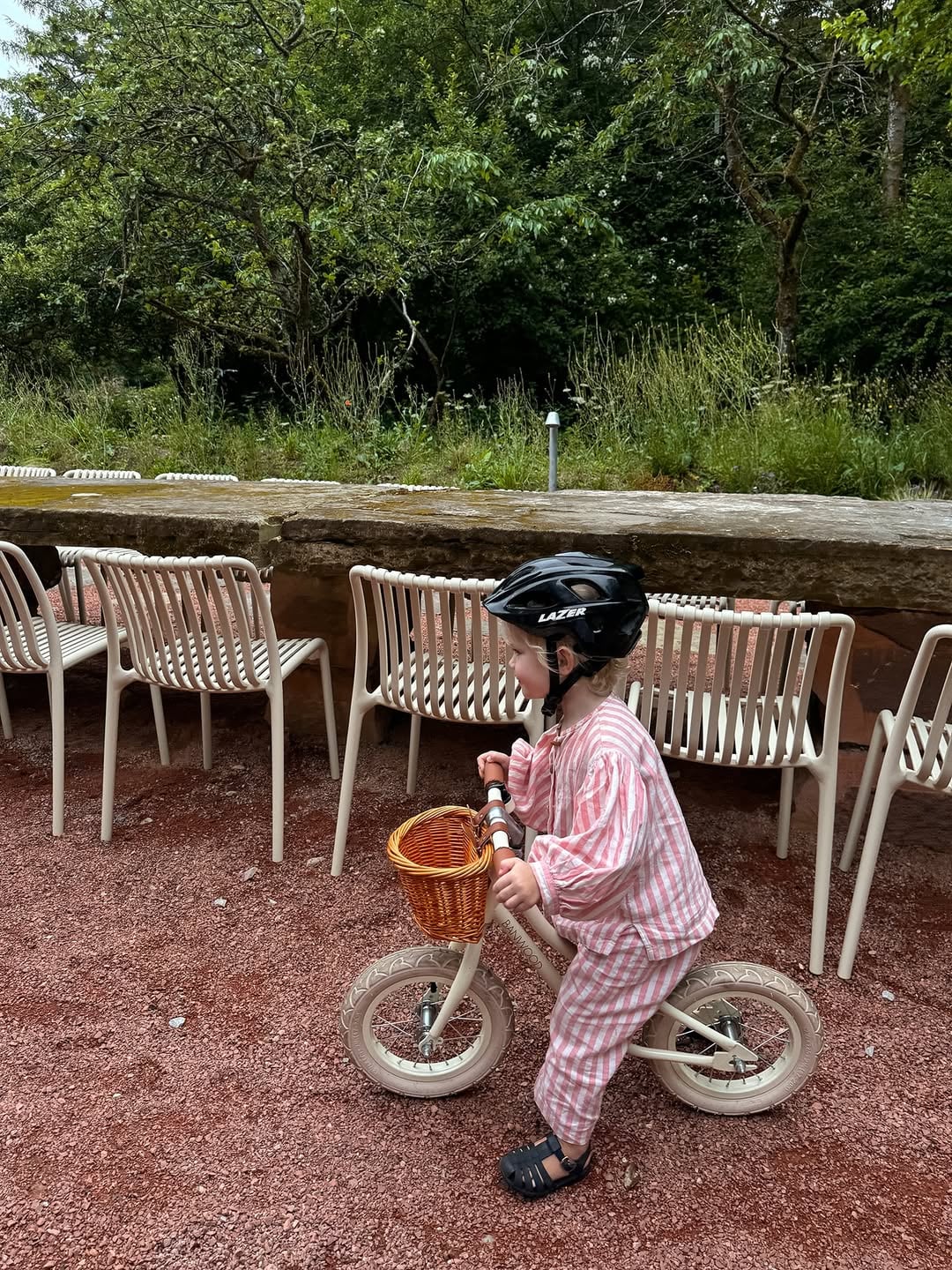 Young child on a balance bike with wicker basket riding on a gravel terrace surrounded by outdoor dining chairs and woodland…