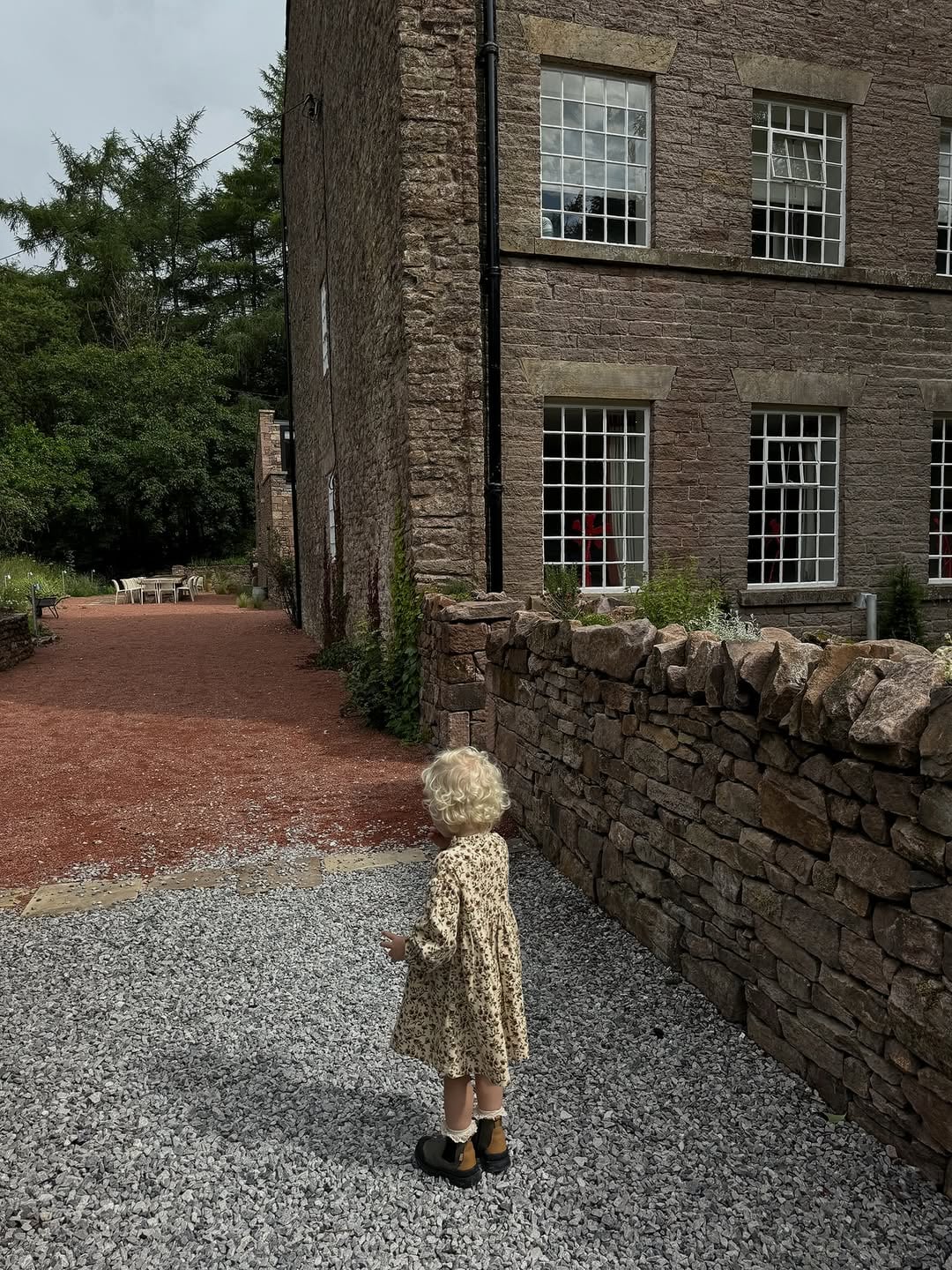 Young child in a floral dress exploring the gravel courtyard beside a grand stone country house with sash windows and dry-st…