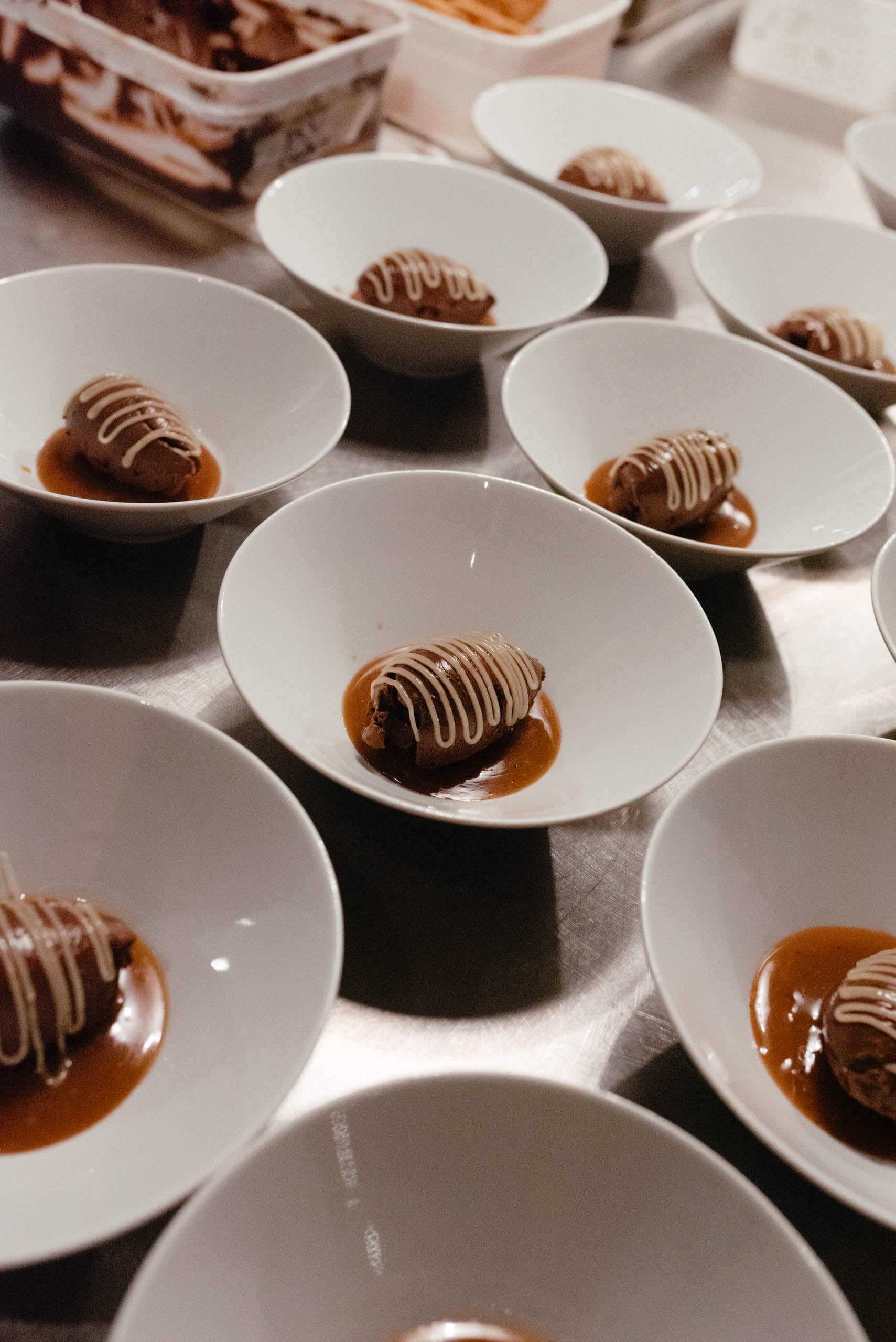 Chocolate desserts with caramel sauce drizzled in white chocolate, plated in white bowls and lined up on a stainless steel k…