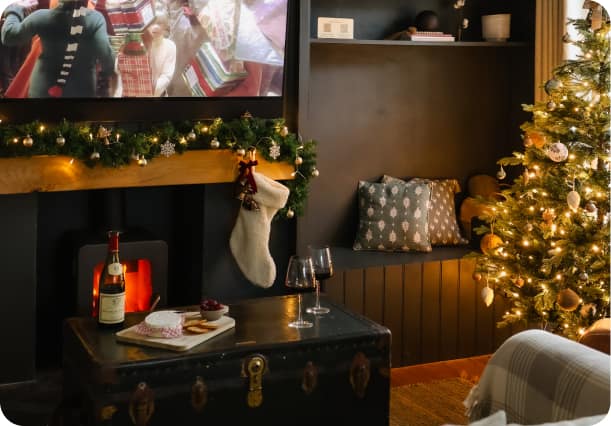 Cosy holiday let living room decorated for Christmas with lit fireplace, garland, stocking, glowing tree, and red wine on a…