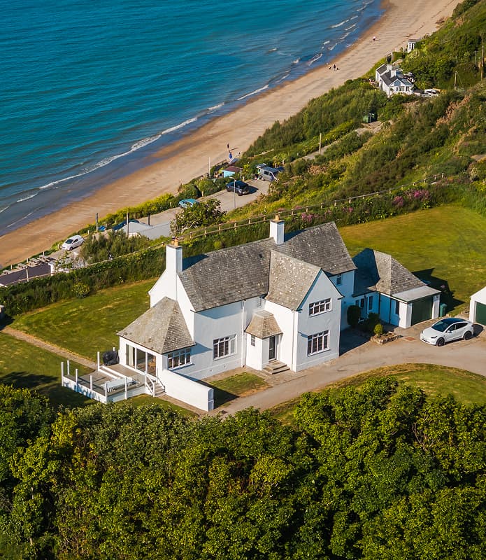 Aerial view of a white clifftop holiday cottage with slate roof and veranda, set above a sandy beach and turquoise sea