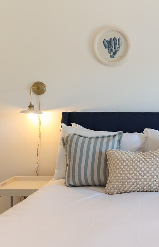 Coastal-style bedroom with navy upholstered headboard, striped blue cushions, brass wall light, and nautical artwork above t…