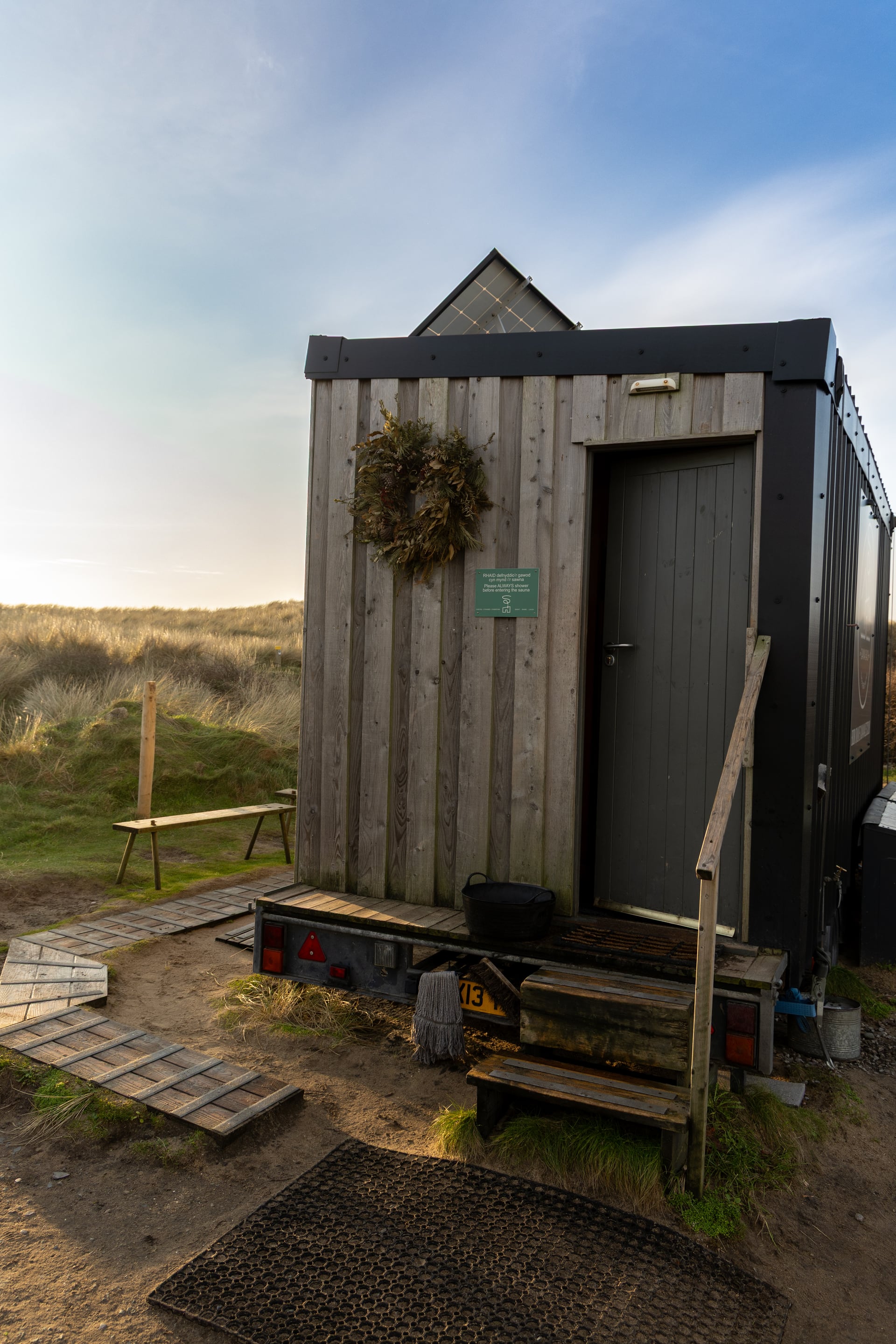 Solar-powered timber sauna cabin with festive wreath on the door, set among coastal sand dunes at golden hour